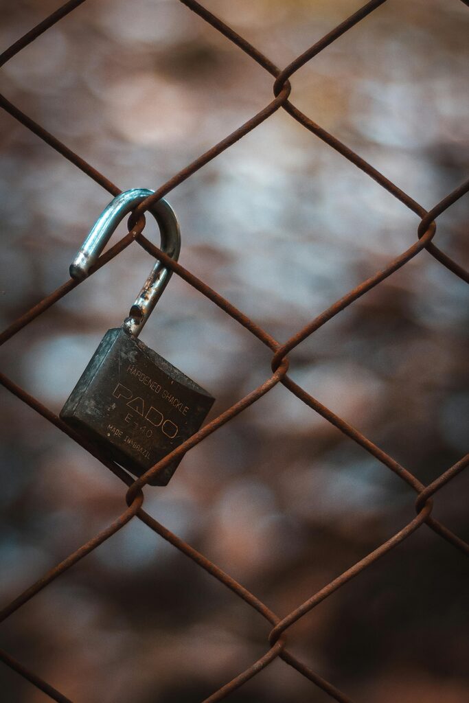 A detailed close-up of a weathered padlock hanging on a chain link fence with blurred background.